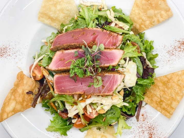 This image depicts a beautiful plate of our Sashimi grade ahi served on top of organic greens, cabbage, carrot, cilantro, grape tomatoes, crisp wonton and almonds complete with an Asian vinaigrette.